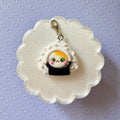 A cute sushi-themed charm featuring a small cartoon cat character peeking out from a white rice bun with black seaweed, displayed on a white scalloped backing card over a light purple patterned background.
