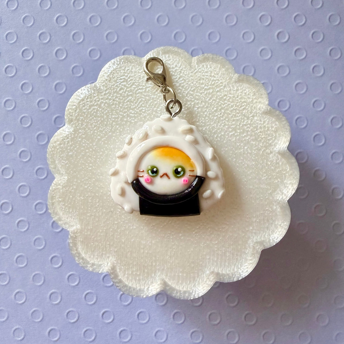 A cute sushi-themed charm featuring a small cartoon cat character peeking out from a white rice bun with black seaweed, displayed on a white scalloped backing card over a light purple patterned background.
